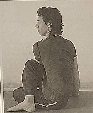 Photograph of a practitioner performing the Ayin twist, left view, seated with one leg folded and torso twisted.