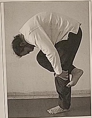 Photograph of a practitioner performing the Kuf posture, bent forward at the waist while balancing on one leg with the other shin supported by the hand.