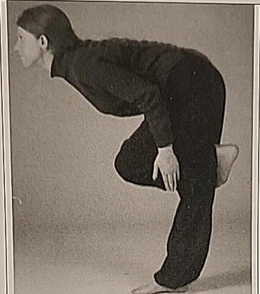 Photograph of a practitioner performing the Resh posture, bent forward at a right angle while balancing on one leg with the other foot tucked behind the knee.