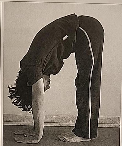 Photograph of a practitioner performing the Tav posture, standing and folded forward at the waist, arms reaching toward the floor.