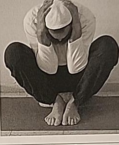Photograph of a practitioner performing the sitting Yud posture from the front, curled forward with knees apart and elbows enclosed by the knees.