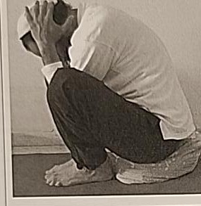 Photograph of a practitioner performing the sitting Yud posture, curled forward with head down and hands covering the ears, side view.